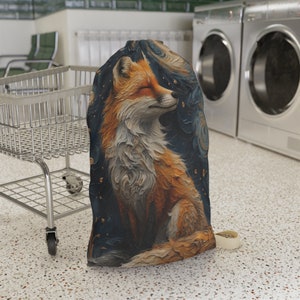 May include: A drawstring laundry bag featuring a detailed illustration of a fox with closed eyes, in shades of orange, white, and brown against a dark blue background with gold accents. The bag has a cream-colored strap.