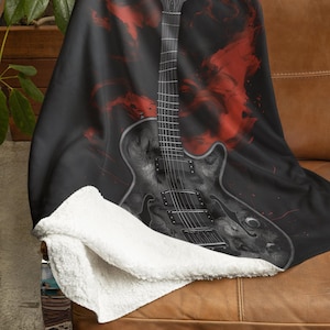 May include: A black electric guitar with red and white paint splatters on a black background. The guitar is on a white and brown throw blanket.