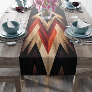 May include: A table setting with a decorative table runner featuring a geometric pattern in shades of red, brown, and beige. The table is set with plates, bowls, silverware, and wine glasses. A vase of pink flowers sits in the center.