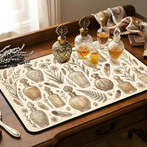 Puede incluir: Alfombrilla de ratón beige con un diseño de botella de perfume vintage, con motivos florales y de plumas. Varias botellas de perfume de vidrio están dispuestas sobre la alfombrilla, junto con otros accesorios, creando una estética clásica y elegante.