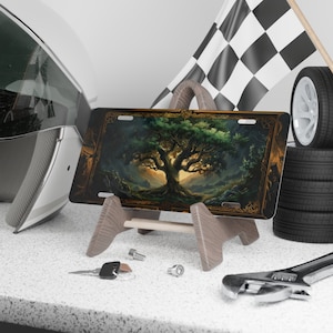 May include: A decorative metal license plate featuring a painting of a large, green tree with a golden frame. The tree is surrounded by a forest and a mountain range in the background. The license plate is on a wooden stand.
