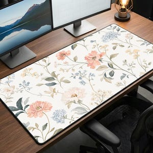 May include: A rectangular desk mat with a floral design. The mat features a cream background with pink, peach, and blue flowers, and green leaves and stems. The mat is on a wooden desk, with a black border.
