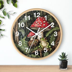Puede incluir: Un reloj redondo de madera con una escena de bosque verde, blanco y rojo. La esfera del reloj tiene números y manecillas blancas, y la escena incluye un hongo rojo y blanco, una rana verde y helechos verdes.