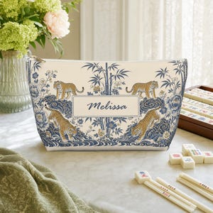 Könnte beinhalten: Ein Reißverschlusstäschchen mit cremefarbenem Hintergrund und einem blau-goldenen Muster aus Tigern, Bambus und Blumen. Ein zentrales rechteckiges Etikett zeigt den Namen Melissa in blauer Schreibschrift. Die Tasche ist konisch geformt.