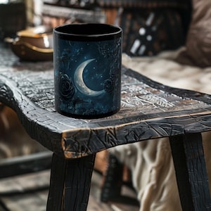 Puede incluir: Taza de café negra con un diseño de luna creciente y estrellas sobre un fondo azul oscuro. El diseño también incluye rosas negras. La taza está sobre una mesa de madera oscura.
