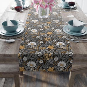 May include: A floral table runner with a dark gray background and a pattern of white and gold flowers and leaves. The runner is placed on a wooden table set for a meal, with plates, bowls, silverware, and wine glasses.