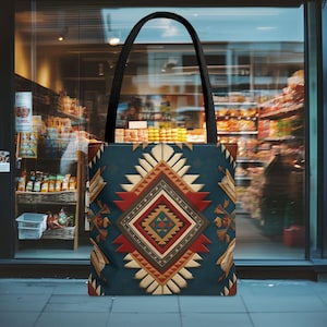 Peut inclure: Un sac fourre-tout avec une anse noire et un fond bleu canard. Le sac présente un motif géométrique dans les tons de rouge, beige et marron. Le motif est centré sur le sac et rappelle l'art amérindien.