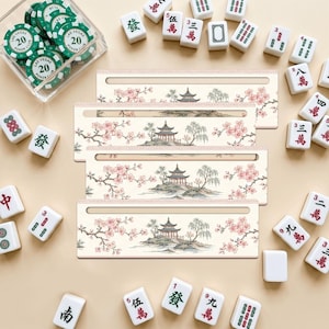 Pode incluir: Quatro suportes retangulares para peças de mahjong com um design impresso de uma pagoda, salgueiros e flores de cerejeira cor-de-rosa. Os suportes estão rodeados por peças de mahjong brancas com caracteres vermelhos e verdes e fichas.