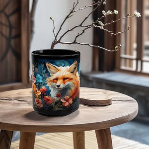 May include: A black ceramic mug with a colorful illustration of a fox surrounded by flowers. The fox has orange fur, a white chest, and black eyes. The flowers are blue, pink, and orange.