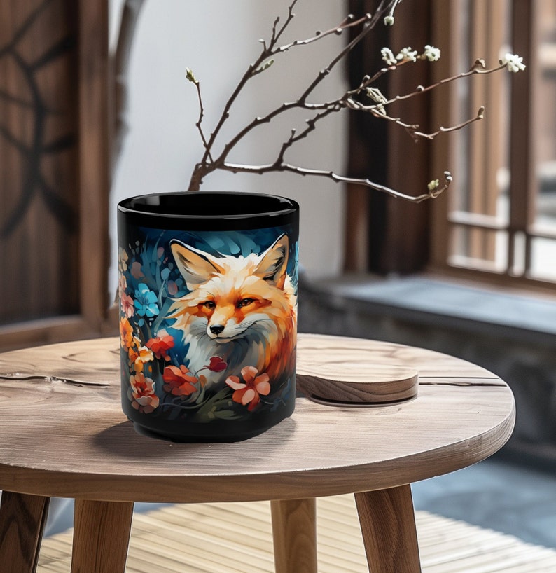 May include: A black ceramic mug with a colorful illustration of a fox surrounded by flowers. The fox has orange fur, a white chest, and black eyes. The flowers are blue, pink, and orange.