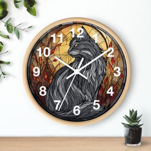 May include: A round wall clock with a light wood frame. The clock face features a stylized gray cat illustration against a stained-glass style background with orange and yellow hues. White numerals mark the hours.