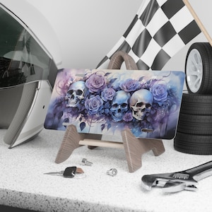 May include: A metal license plate featuring a watercolor design of three skulls surrounded by purple roses and vines. The license plate is on a wooden stand.