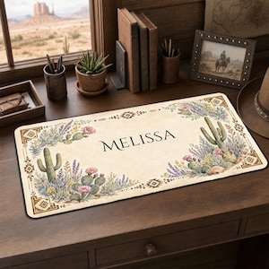 May include: A rectangular desk mat with a cream-colored background featuring a desert-themed border of cacti, succulents, and pink flowers. The name MELISSA is printed in black serif font in the center, framed by geometric corner patterns.