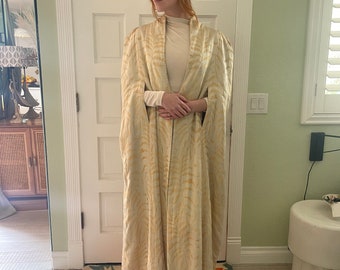May include: A long, cream-colored cape with a subtle leaf pattern. The cape has a high collar and long sleeves. The model is wearing a white turtleneck and a white fur hat.