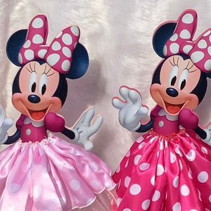 Op de afbeelding: Drie kartonnen uitknipsels van Minnie Mouse die verschillende gekleurde jurken dragen. De Minnie Mouse links draagt een roze jurk met witte stippen. De Minnie Mouse in het midden draagt een felroze jurk met witte stippen. De Minnie Mouse rechts draagt een rode jurk met witte stippen.