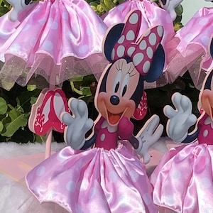 May include: A set of four pink and white Minnie Mouse centerpieces. Each centerpiece features a Minnie Mouse character with a pink polka dot dress and a white bow. The centerpieces are perfect for a Minnie Mouse themed party.