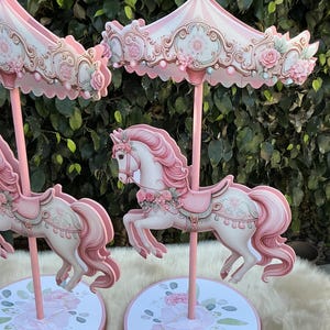 May include: Pink carousel horse decorations with floral details. The carousel horses have pink manes and tails, and are decorated with pink roses. The carousel tops are striped pink and white, with floral and scroll details. The bases are round and decorated with pink roses.