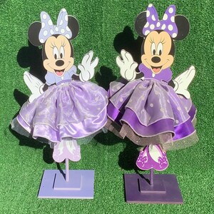 May include: Two purple Minnie Mouse centerpieces with white polka dots and a purple dress. Each centerpiece has a purple base and is standing on a green surface.