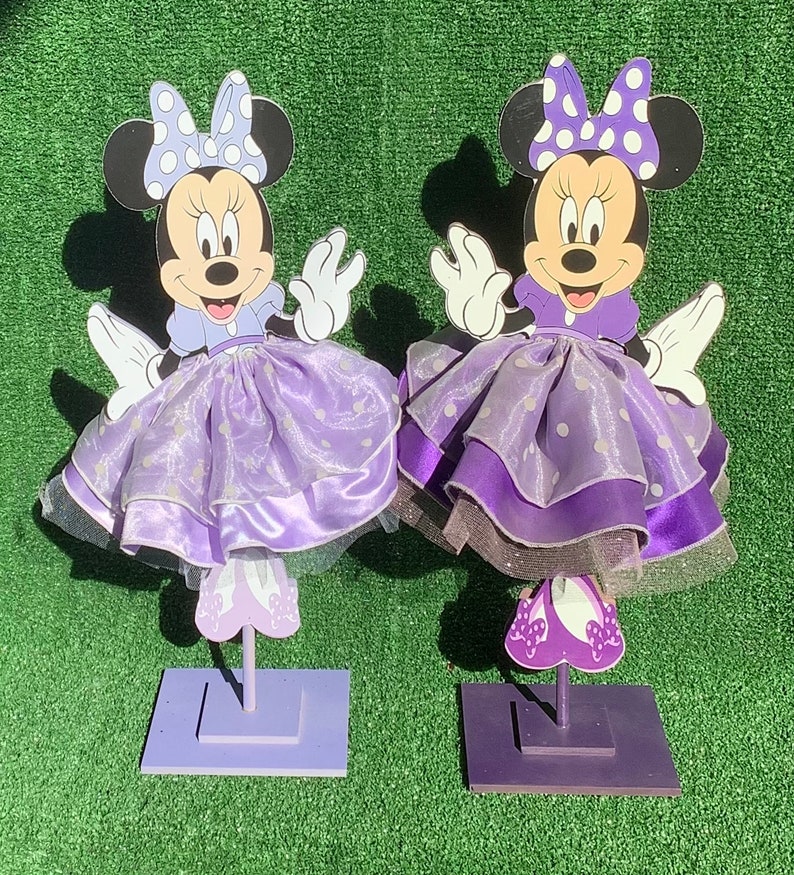 May include: Two purple Minnie Mouse centerpieces with white polka dots and a purple dress. Each centerpiece has a purple base and is standing on a green surface.