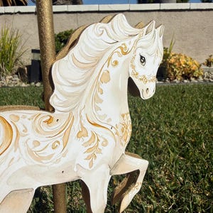 May include: A white carousel horse with gold accents and decorative floral patterns. The horse has a flowing mane and tail, and is mounted on a gold pole. The carousel horse is set against a backdrop of green grass and a blue sky.
