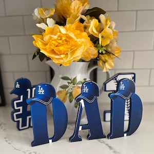 May include: Blue wooden letters spelling "DAD" with a baseball cap on top of each letter. The caps are blue with a white "LA" logo. The letters are sitting on a white surface.