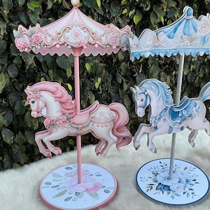 May include: Two carousel horse decorations. One is pink with floral accents, the other is blue. Both have decorative canopies and sit on round bases with floral patterns. The background is a lush green.