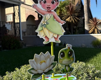 Könnte beinhalten: Ein Set aus Gartendekorationen mit einer Cartoon-Prinzessin in einem grünen Kleid, einer weißen Lotusblume und einem Cartoon-Frosch. Die Prinzessin winkt und der Frosch trägt eine Krone. Die Dekorationen stehen auf gelben Stangen in einem grünen Busch.