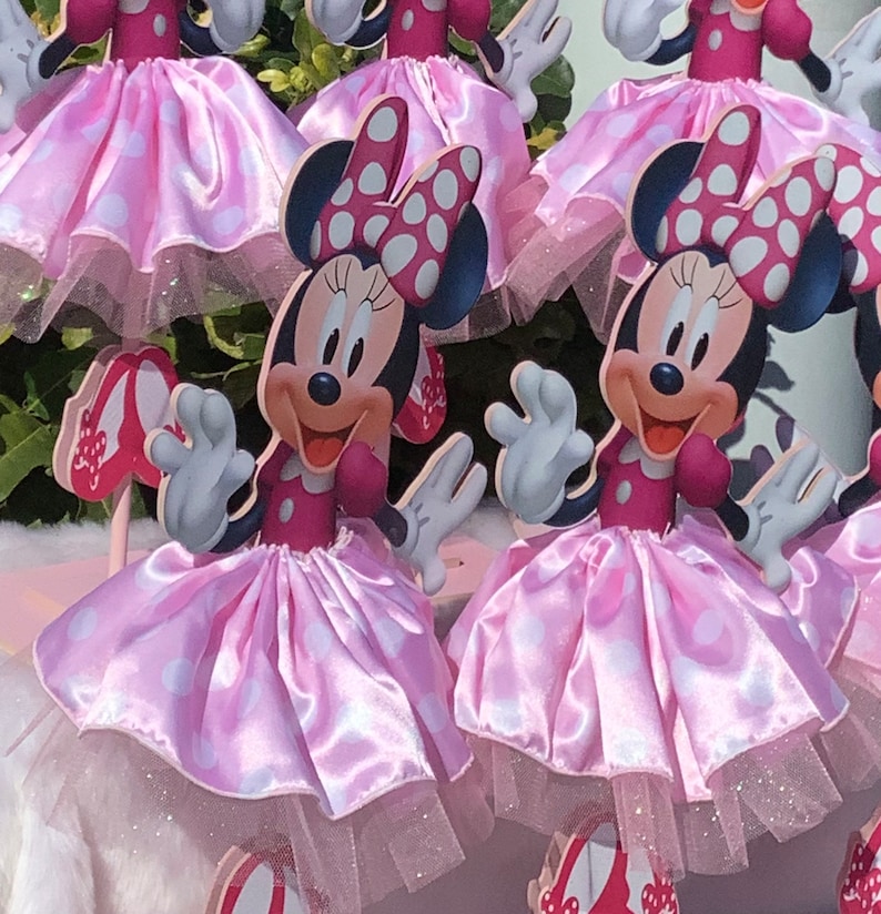 May include: Pink and white Minnie Mouse centerpieces with a pink tulle skirt and white polka dots. Each centerpiece features a Minnie Mouse character with a red bow and white polka dots.