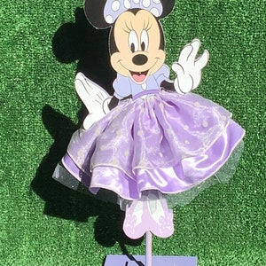 May include: A purple and white Minnie Mouse character cutout with a purple dress and white polka dots. The character is standing on a purple stand.