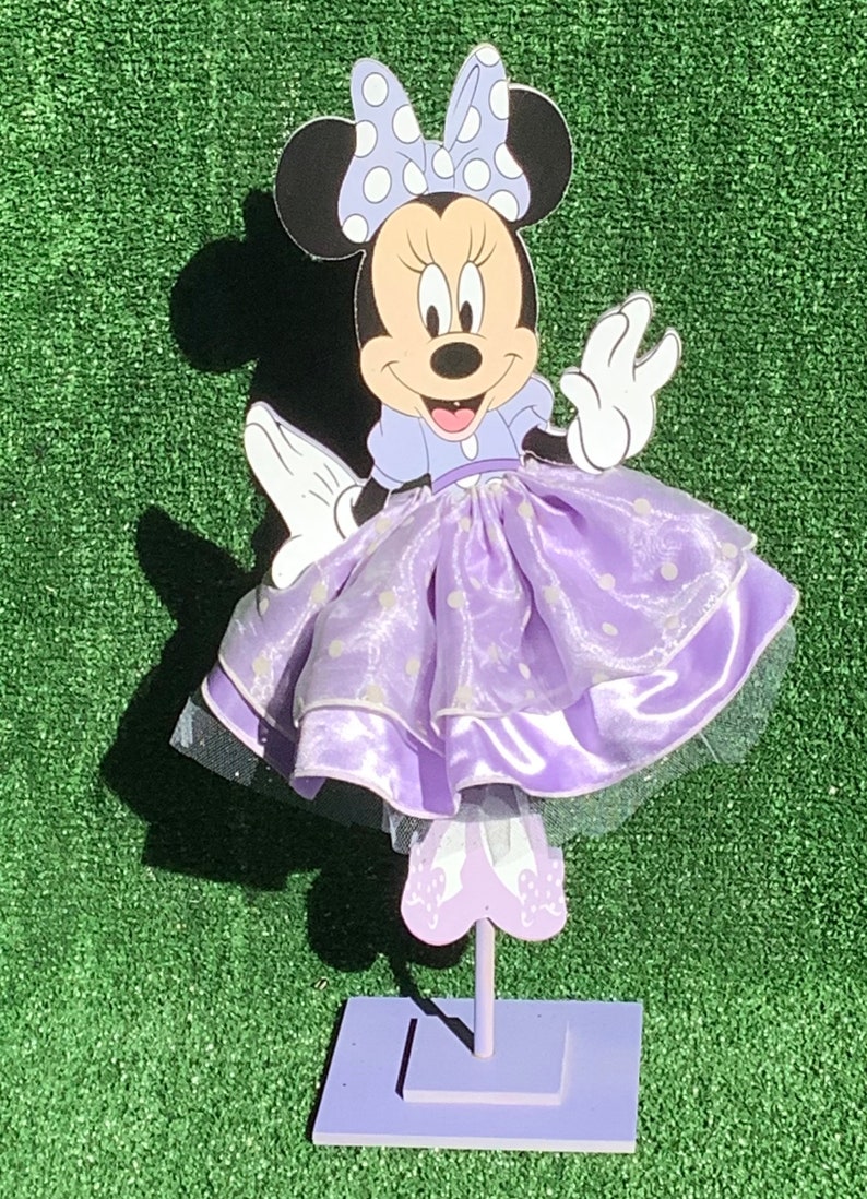 May include: A purple and white Minnie Mouse character cutout with a purple dress and white polka dots. The character is standing on a purple stand.