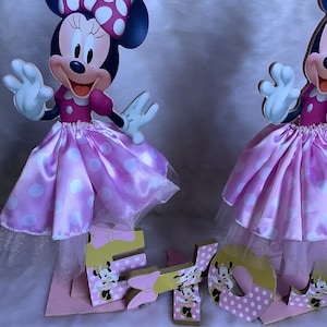 May include: Two pink and white Minnie Mouse centerpieces with gold accents. Each centerpiece features a Minnie Mouse character with a pink polka dot skirt and a white tulle overlay. The centerpieces are decorated with gold glitter and have the letters "E" and "L" on the base.