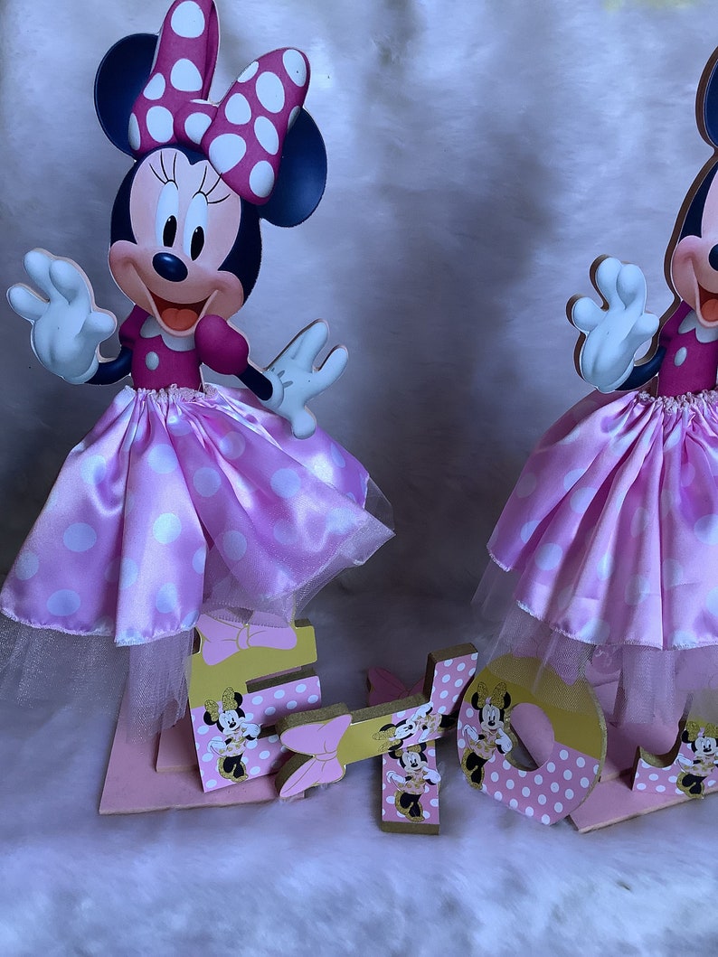 May include: Two pink and white Minnie Mouse centerpieces with gold accents. Each centerpiece features a Minnie Mouse character with a pink polka dot skirt and a white tulle overlay. The centerpieces are decorated with gold glitter and have the letters "E" and "L" on the base.