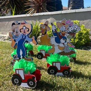 以下が含まれることがあります： ミッキーマウス、ドナルドダック、グーフィー、プルートの4つのカートゥーンキャラクターのセンターピース。各キャラクターは農場をテーマにした服装をし、緑の草が生えた赤いトラクターに乗っています。