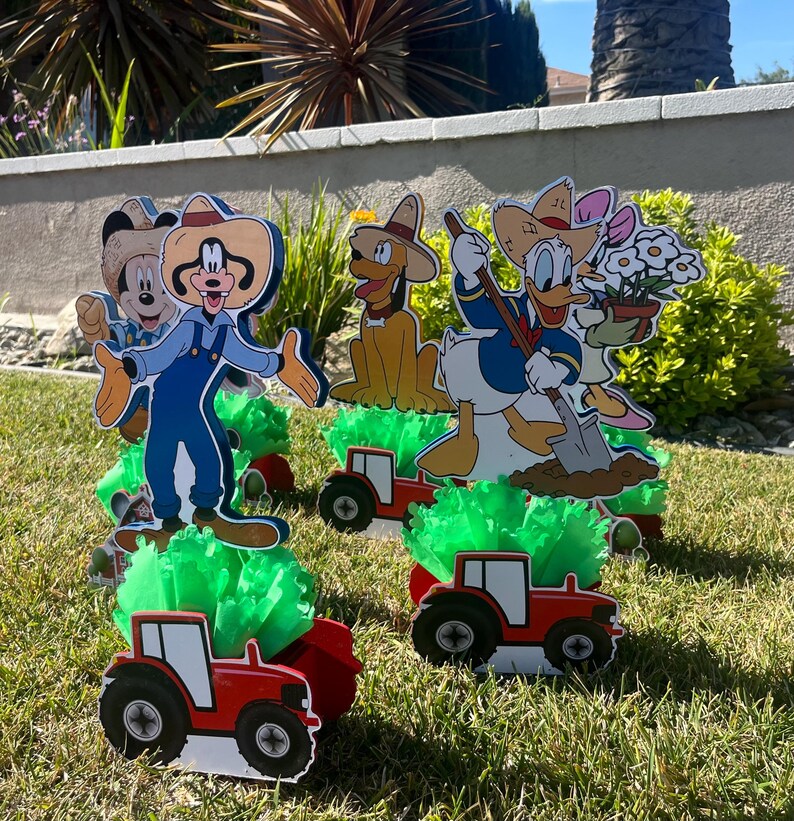 以下が含まれることがあります： ミッキーマウス、ドナルドダック、グーフィー、プルートの4つのカートゥーンキャラクターのセンターピース。各キャラクターは農場をテーマにした服装をし、緑の草が生えた赤いトラクターに乗っています。