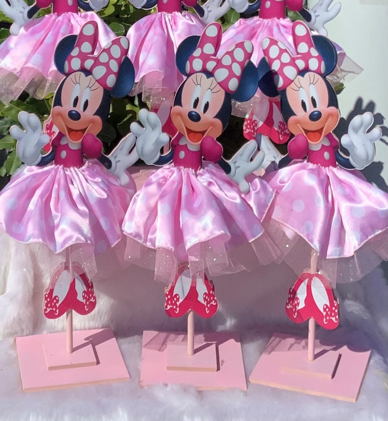 May include: Set of four pink and white Minnie Mouse centerpieces. Each centerpiece features a Minnie Mouse character with a pink and white polka dot dress and red shoes. The centerpieces are on pink square bases.