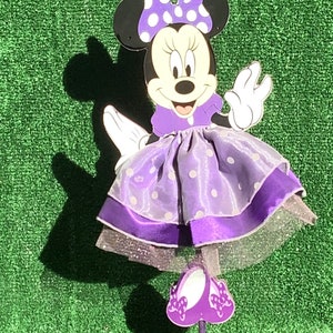 May include: Purple Minnie Mouse figurine with a white polka dot dress and bow. The figurine is standing on a purple base.