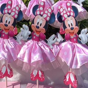 May include: Three pink and white Minnie Mouse centerpieces with red shoes and polka dot bows. The centerpieces are made of cardboard and have a tulle skirt.
