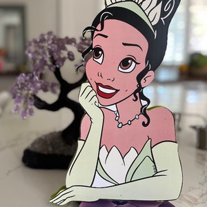 Op de afbeelding: Een houten beeldje van Prinses Tiana uit Disney's The Princess and the Frog. Ze draagt een groene jurk met een witte bovenkant en een kroon. Ze glimlacht en heeft haar hand op haar kin. Het beeldje staat op een paarse bloem.