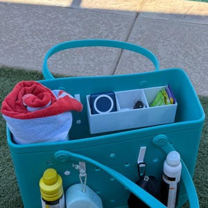 May include: A teal rubber tote bag with perforated sides and two handles. The interior contains a white divided organizer, a rolled red and white towel, and small items. The exterior features a yellow bottle, a white case, and a white water bottle.