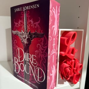 May include: A hardcover book titled "Dire Bound" by Sable Sorensen, featuring a sword design on a burgundy cover. A red "Love" sign and a rose are in a white shelf divider.