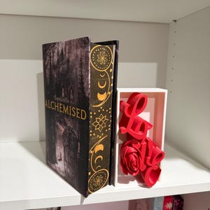 May include: A hardcover book titled "Alchemised" with a dark, gothic cover design, displayed on a white shelf. The spine is black with gold floral and crescent moon patterns. A red "Love" sign and rose are in a white box.