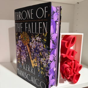 May include: A hardback book titled "Throne of the Fallen" by Kerri Maniscalco, with a gold key and purple floral design on the cover. The book has purple floral edges and is displayed with a red "Love" decoration.