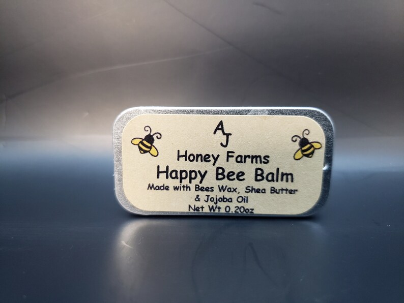 May include: A rectangular tin of Happy Bee Balm from Honey Farms. The label features a yellow and black bee graphic and text that reads "Happy Bee Balm" and "Net Wt 0.20oz". The balm is made with Bees Wax, Shea Butter, and Jojoba Oil.