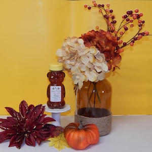 May include: A glass jar with a brown, rustic rope tied around the top, filled with a bouquet of white and orange flowers and red berries. A small bottle of honey with a bear-shaped bottle and a label that reads "A.J. Honey Farm Raw Honey" is on a white pedestal in front of the jar. A small orange pumpkin sits on the table in front of the jar.