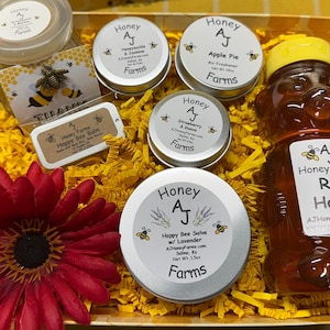 May include: A gift box filled with honey products from Honey AJ Farms. The box contains a jar of raw honey, a lip balm, and four small jars of honey infused with different flavors, including apple pie, strawberry & guava, honeysuckle & jasmine, and lavender. The box is filled with yellow shredded paper and a red flower.