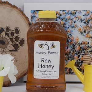 May include: A clear glass bottle of raw honey with a yellow lid. The label says "A J Honey Farms" and "Raw Honey". The bottle is sitting on a wooden surface with a honeycomb design.