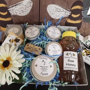 May include: A gift box filled with honey products from AJ Honey Farms. The box contains a jar of raw honey, a jar of honey soap, a jar of honey salve, a jar of honey lip balm, and a small wooden bee-shaped honey dipper. The box is decorated with a bee-themed design and the words "bee kind".