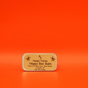 May include: A rectangular tin of Happy Bee Balm from Honey Farms, with text and bee illustrations on a tan label. The label states the balm is made with beeswax, shea butter, and jojoba oil, with a net weight of 0.20 oz. The background is a solid orange.