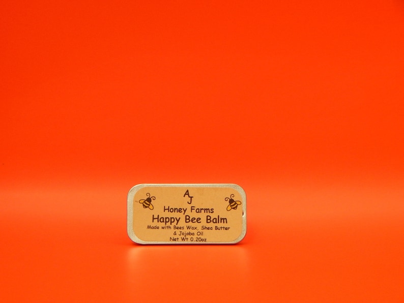 May include: A rectangular tin of Happy Bee Balm from Honey Farms, with text and bee illustrations on a tan label. The label states the balm is made with beeswax, shea butter, and jojoba oil, with a net weight of 0.20 oz. The background is a solid orange.