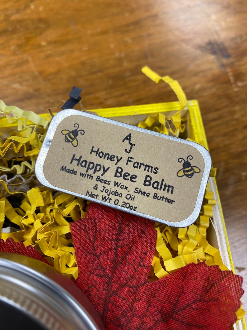 May include: A small, rectangular tin of Happy Bee Balm sits in a yellow box filled with yellow paper shreds and a red leaf. The tin has a brown label with the text "Happy Bee Balm" and "Net Wt 0.20oz". Two bee illustrations are on the label.